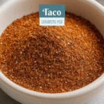 A small white bowl of homemade taco seasoning surrounded by warm spices like chili powder, paprika, and cumin on a wooden counter.